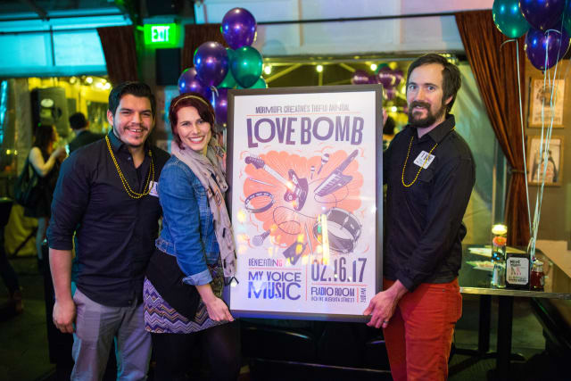 Three individuals posing with a colorful poster for the Love Bomb event, featuring musical instruments and the text "Love Bomb" and "benefiting My Voice Music," with festive balloons in the background.